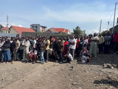 A group of approximately 50 people are gathered on a dirt road in the city of Bukavu, Democratic Republic of Congo. They are standing and sitting in a loose formation, facing toward the camera. The majority of the people are men, but there are also several women and a few children present. The people are mostly dressed in casual clothing. Some are wearing hats. The surrounding environment is urban, with buildings visible in the background. There is a single utility pole and power lines in the image. There is a small child sitting on the lap of a woman wearing a floral patterned dress. A man in a long, loose-fitting robe stands in the center of the group with his arms crossed. The people in the image are likely residents of the Bukavu area and appear to be participating in some kind of informal gathering.