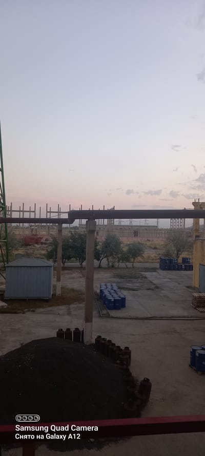 In Levakant, Tajikistan, this image captures an outdoor industrial setting during a dusk or dawn. A large pile of dark, granular material, possibly coal or fertilizer, is visible in the foreground, bordered by a red barrier and lined with numerous cylindrical objects resembling propane tanks. In the middle ground, a row of blue barrels is neatly stacked, and a small, grey metal storage unit is situated to the left. Further back, trees with sparse foliage dot the landscape, and the skeletal remains of unfinished structures rise against the pale sky. In the distance, electrical pylons suggest the presence of a power infrastructure. The overall scene evokes a sense of ongoing or halted industrial activity in an arid or semi-arid environment.