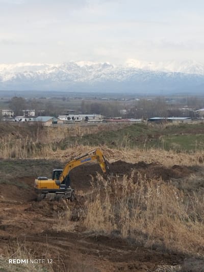 This image depicts an outdoor panoramic scene in Nurli zamin, Uzbekistan, under an overcast sky. In the foreground and midground, a construction site is active. A yellow-and-black XCMG XE150W wheeled excavator is situated on disturbed, dark brown earth, with its articulated boom and bucket engaged with a pile of excavated soil. Tall, dried, light brown reeds or grasses surround the immediate excavation area. Beyond this, in the midground, a low-density settlement is visible, characterized by numerous single-story or low-rise buildings with light-colored facades and various roof structures. Sparse, bare deciduous trees are interspersed among the buildings. The distant background is dominated by a continuous range of rugged, snow-capped mountains extending across the horizon, silhouetted against a uniform light grey, cloudy sky. No human figures are discernible.