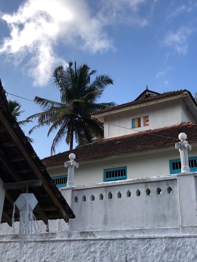 The image shows a white building with a tiled roof and a pale blue sky with some clouds. There are palm trees in the background. The building has a decorative wall with pointed openings. A white paper lantern hangs from the eaves of another roofed structure on the left. The building has blue-framed windows and a smaller window with colored panels on the upper floor. The overall scene is tranquil and suggests a warm climate. This building is located in Pilana, Sri Lanka.