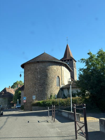 The image depicts a cylindrical stone building with a conical tiled roof, possibly a church or a historical tower, set against a clear blue sky. A pointed, brown-tiled roof with a weathervane crowns the structure. The stone walls are rough-hewn and appear aged. A small, arched window with a darkened pane is visible on the rounded facade. A decorative wrought-iron lantern hangs from a bracket below the roofline.

To the right of the building, a paved road runs parallel to a low stone wall, topped with a metal railing. Dense green foliage and trees are visible behind the wall. Several road signs are present: one blue and white sign with symbols, and another circular sign with a red border and a horizontal white bar, indicating a prohibition, possibly "no entry" or "no vehicles." Further down the road, a white van is parked.

The scene suggests a daytime setting, with bright sunlight casting shadows on the road. The weather appears clear and sunny. The architecture and signage hint at a location in a village or town, consistent with the provided context of Villages du Lac de Paladru, France. There are no people visible in the image, and the overall impression is one of a quiet, historical European setting. The orientation of the image is rotated 90 degrees clockwise.