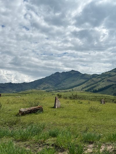 The image depicts a serene landscape featuring rolling green hills and mountains under a cloudy sky. In the foreground, several large, weathered stones are scattered across a grassy meadow. One prominent stone lies horizontally, covered in moss, while another stands vertically to its right. Further into the field, more stones are visible, including a tall, triangular stone. The background is dominated by the majestic, layered slopes of the mountains, dotted with sparse trees. The overall scene suggests a natural, possibly ancient, setting. The location is identified as Kazanovka, Russia. There are no people or animals visible in the frame.