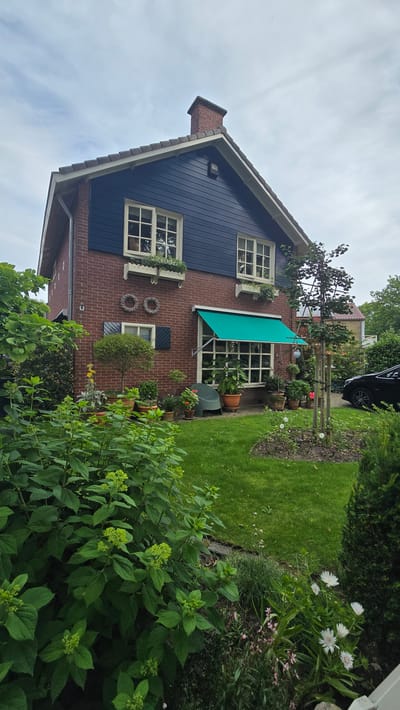 The image shows a brick house with dark blue siding on the upper level, located in Zeddam, Netherlands. A green awning extends over a large window on the first floor. The front yard is landscaped with a lush green lawn, various plants, and potted flowers. A dark-colored car is parked in the driveway to the right. The sky is overcast.