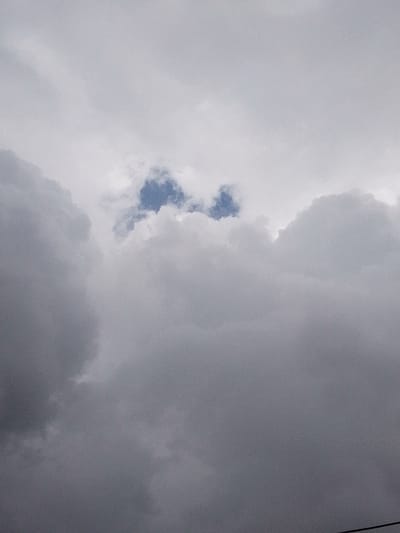 The image displays an overcast sky above Nachu ward, Kenya. The frame is dominated by multiple layers of grey clouds, exhibiting variations in density and luminance, ranging from dark grey, particularly in the lower left, to lighter white-grey across the upper and central areas. Two small, irregular patches of light blue sky are visible through breaks in the cloud formation in the upper-central portion of the image. No human figures, animals, terrestrial structures, or discernible natural land features are present. A faint, thin dark line, consistent with a utility wire or cable, is visible extending diagonally from the bottom-right corner of the frame.