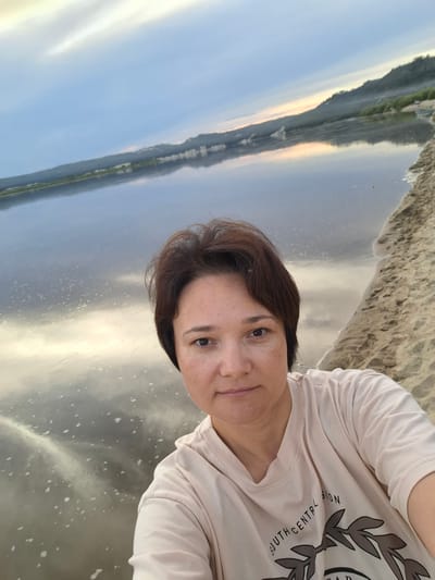 In the city of Malaya Sazanka, Russia, a woman of Asian descent with short, dark brown hair and fair skin with freckles is taking a selfie on a sandy beach next to a large body of water. She is wearing a light-colored t-shirt with a graphic print that reads "SOUTH CENTRAL UNION." The water reflects the sky, which is filled with a mixture of blue and white clouds, with a hint of orange and yellow on the horizon, suggesting either sunrise or sunset. In the background, a tree-lined hillside rises from the water. The overall scene is serene and appears to be a natural landscape.