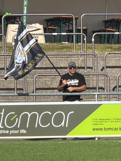 A medium shot, taken in daylight, presents a man standing behind a silver-toned railing, seemingly at a sporting event, given the green patch resembling a field. The man is dark-haired, wearing a cap and sunglasses, and a dark t-shirt adorned with a graphic. He is partially obscured by the railing, but is positioned next to a black, gold, and white flag.
To the right of the man is a large sign advertising "tomcar" with the tagline "drive your life." The sign includes business contact information in Luxembourg (9, ZAC Klengbousbierg 1-7795 Bissen; T+352 26887395; info@tomcar.lu; www.tomcar.lu). The background shows outdoor seating, including green benches and wooden tables. The setting suggests an outdoor venue possibly in Fentange, Luxembourg, based on the address listed.