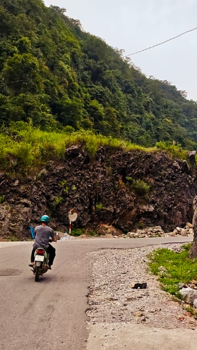 A motorcyclist with a blue helmet is traveling on a paved road in the Cán Tỷ Commune, Vietnam. The road is surrounded by a steep, rocky hillside covered in lush, green vegetation. A convex mirror is mounted on a pole along the roadside. The road surface appears to be patched with gravel and is in a state of disrepair.