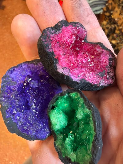 A close-up image taken in Arucas, Spain, depicts an individual's hand holding three distinct, colorful geode formations. The hand, exhibiting visible skin texture and minor dark smudges on the fingers, serves as the primary support for the objects. The uppermost geode presents a vibrant fuchsia or hot pink crystalline interior, encased by a rough, dark, charcoal-like exterior shell. To the lower left, a second geode displays a brilliant purple crystalline structure within a similar dark shell. The third geode, situated to the lower right, features vivid green crystals also contained within a dark, rugged outer layer. All three geodes exhibit crystalline facets that reflect light. The background is blurred, showing warm, earthy tones consistent with an indoor environment, possibly a wooden surface. No other individuals or specific actions are discernible beyond the hand holding the objects.