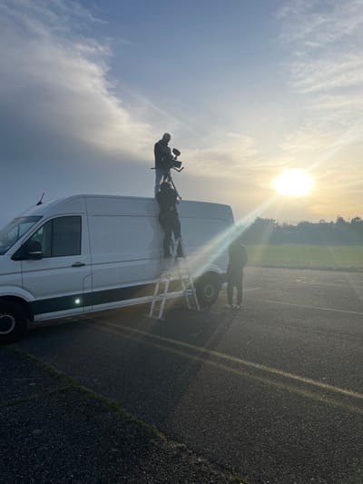 An outdoor scene in an unspecified area of Reventin-Vaugris, France, depicts three individuals engaged in a filming or photography setup at either sunrise or sunset, indicated by a low sun and prominent lens flare. A white commercial van is centrally positioned on a dark asphalt surface with faded yellow ground markings. One individual, dressed in dark clothing and a cap, stands on the van's roof, operating a video camera mounted on a tripod, directed towards the sun-drenched horizon. A second individual, also in dark clothing, is positioned on a white step ladder propped against the passenger side of the van, appearing to support the setup or ascend to the roof. A third individual, wearing dark attire and sneakers, stands on the asphalt adjacent to the van, observing the activity. The environment includes a grassy edge in the foreground and a hazy horizon with distant vegetation and possibly structures under a partly cloudy sky.