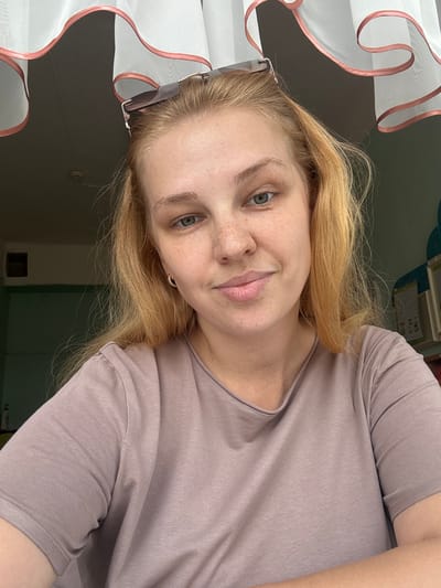 A young Caucasian woman with blonde hair and freckles is pictured in a selfie. She is wearing a taupe t-shirt and has sunglasses perched on her head. The background appears to be an indoor setting, possibly a home, with a window covered by sheer white curtains with pink trim. The lighting suggests it is daytime. The setting is not identifiable as being specifically in Vakhrushovo, Russia.