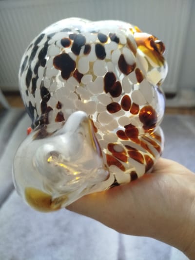 A hand holds a decorative glass owl figurine. The owl is clear glass with irregular white, dark brown, and amber speckles. The figurine is held in a person's right hand, which is cupped from below. The background is blurred and appears to be an indoor setting with a radiator visible on the far left. The image does not depict any location in Offenbach am Main, Germany.