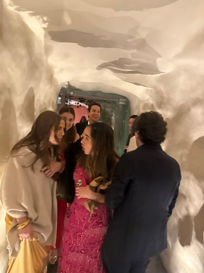 Inside a bright space with textured walls, a group poses. A man in a dark blue jacket stands tall, partially obscuring a woman in a bright pink, sequined dress holding tan shoes. Below them, two women stand closer to the camera, one in a tan sweater and another behind her. A man smiles in the background. The scene appears to be captured in a mirror, reflecting the group and their surroundings.