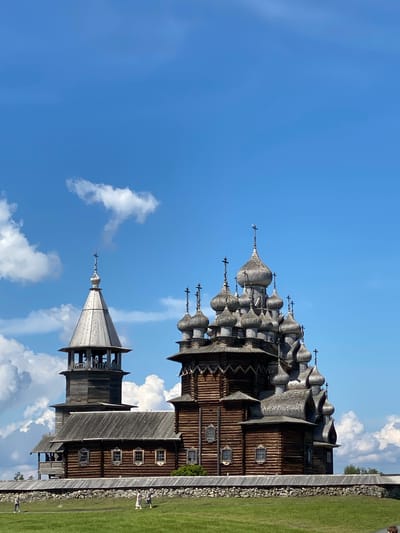 The image depicts the Kizhi Pogost in Boyarshchina, Russia, a UNESCO World Heritage site. The focal point is the intricately designed wooden Church of the Transfiguration, characterized by its numerous onion domes and traditional Russian architecture. Adjacent to it stands a wooden bell tower. Both structures are constructed primarily of weathered wood, with visible log construction. They are situated on a grassy knoll, bordered by a low stone wall. The sky above is a vibrant blue, dotted with scattered white cumulus clouds. In the foreground, a few individuals are walking across the green lawn.