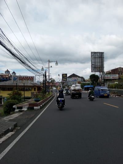 The image presents a street scene in Taohom, Laos, under an overcast sky. A multi-lane asphalt road occupies the foreground and mid-ground, delineated by a solid white line on the left and a solid yellow line separating lanes further right. On the far left, a paved sidewalk abuts a stepped concrete barrier, behind which is a raised planter section containing foliage.
Traffic moves from the foreground towards the background. A motorcyclist wearing dark attire and a white helmet operates a dark scooter with a visible blue light on its rear, positioned in the left lane. Ahead and to the right, a dark pickup truck is heavily laden with multiple large white sacks, piled significantly above its cab. To the right of the truck, a second motorcyclist in dark clothing and a dark helmet rides a dark scooter, followed by a blue utility van. Additional vehicles, including cars and a yellow vehicle, are visible further down the road in the background.
Along the left side of the street, a yellow building with a dark roof is prominent, featuring a large sign that reads "IDAMAN HOTEL COTTAGE" in white letters with accompanying Indonesian text. Partially visible behind this is a building with a blue dome-like roof structure. Numerous overhead utility wires form a dense cluster across the upper left portion of the frame, suspended by poles. These poles also feature decorative streetlights with lantern-style fixtures.
On the right side of the thoroughfare, a large, empty metal billboard structure stands tall. Further down on the right, a sign displaying "AYAM GORENG PANAS INDAH" (Fried Chicken Hot Beautiful) is visible on a building. Other commercial buildings with varying architectural styles line the street. Green foliage and shrubs are present along the roadside. No pedestrians are distinctly visible.