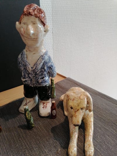 This is a close-up, eye-level, indoor shot of a collection of ceramic figurines. In the center-left, a roughly formed ceramic figurine of a person stands upright. The figurine has a pale skin tone with reddish-brown hair and appears to be wearing a blue and white patterned jacket over black pants and light-colored shoes. The figurine is holding a green bottle in its right hand. To the right of the standing figurine, a pale yellow ceramic dog figurine lies down. The dog has brown speckles and appears to be looking forward. In the foreground, between the two main figurines, are two small ceramic bottles, one green and one reddish-brown. The figurines are placed on a dark grey ribbed mat. Behind the figurines is a wooden surface, and further back, a white textured wall. The scene is lit with artificial light. The location is not specified in the image.