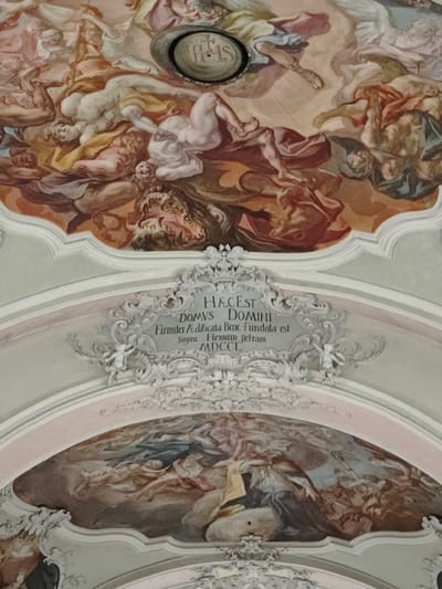 The image shows a close-up of a frescoed ceiling in the Filzen, Austria area, likely within a church or public building. The fresco depicts a dramatic scene with numerous figures, including cherubic putti, angels, and human-like figures. The style suggests Baroque or Rococo art, characterized by swirling drapery, dynamic poses, and a sense of movement. In the upper portion of the image, a circular medallion with the inscription "IHS" is visible. Below this, a decorative cartouche contains Latin text. The text reads: "HÆC EST DOMVS DOMINI Firmiter Ædificata bene Fundata est Supra Firmam æt [sic] MDCCLXXXI". This translates to "This is the House of the Lord, firmly built and well founded, above the firmament, 1781." The lower part of the image reveals another section of the fresco, continuing the theme of heavenly or allegorical figures. The colors are muted, with dominant tones of cream, gold, brown, and pale blue, creating a sense of grandeur and spirituality.