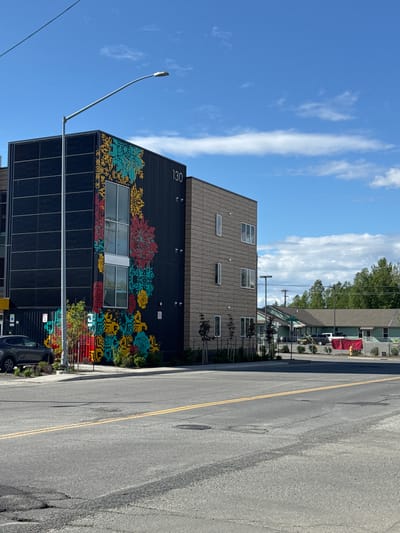 This is a street-level, eye-level, full shot of a modern building with a colorful mural on its side, located in Anchorage, Alaska. The building is dark grey with a section covered in wood-like siding. The mural features stylized floral patterns in yellow, teal, red, and black. The building has several windows, and the number "130" is visible on the dark grey section. A street lamp with a curved arm extends over the building. In front of the building, there is a dark grey car parked, and some landscaping with small trees and bushes. The foreground shows a paved road with yellow lane markings and some patches of wear. In the background, across the street, are several low-rise buildings, one of which is green, and some trees line the horizon under a blue sky with scattered white clouds. There are no people visible in the image.