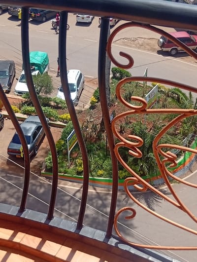 The image provides an elevated, oblique perspective of a street scene in Murang'a, Kenya, observed from behind a dark-grey and copper-toned ornamental metal balcony railing. The railing, featuring scrolled decorative elements, casts distinct shadows onto a wooden plank floor below.

The visible environment consists of an asphalt road traversing diagonally, marked with white solid and dashed lane lines. Numerous vehicles are present; a dark-colored sedan, a white sedan, and a person on a motorcycle are on the main road in the upper frame. A red/maroon SUV with an externally mounted spare tire is parked on a dirt shoulder to the upper right.

Adjacent to the road, on a paved or unpaved parking area, are several parked vehicles: a dark sedan, a white pickup truck with a green tarp covering its cargo bed, a silver/white station wagon, and a partially obscured grey sedan. A person wearing red pants and a dark top is standing near the silver/white station wagon, next to a parked motorcycle. Another individual in a yellow top is visible near the dark sedan in the upper left.

Landscaped sections containing various green shrubs, small trees, and succulent plants are interspersed between the road and parking areas, often delineated by concrete curbs painted green and orange. A white rectangular sign with blue lettering "OPTICAL" is mounted on a pole within one of these planted areas. The bright daylight casting sharp shadows, including those of unseen palm trees on the road and greenery, indicates clear weather. The road surface exhibits signs of moderate use, including minor unevenness and small worn patches.