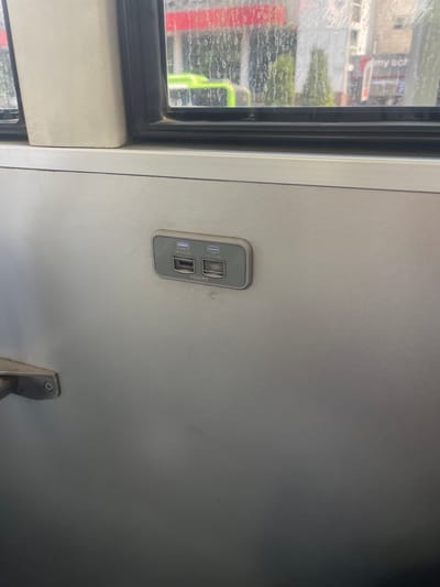 The image shows a close-up view of a wall inside a vehicle, possibly a bus or train. Mounted on the wall is a grey dual USB charging port. The port has two USB-A sockets, each labeled with "5V=2.4A". Above the sockets, there are small indicator lights, one of which appears to be lit blue. The wall itself is a light grey, metallic-looking material. To the left of the charging port, a silver metal handle is partially visible, attached to the wall with a screw. The background of the image shows a window with condensation or water streaks on the glass. Through the window, a blurry view of an urban environment can be seen, with buildings and a bright green object, possibly a vehicle. The context of Kashgar mahalla, Uzbekistan, is not visually discernible from the image.