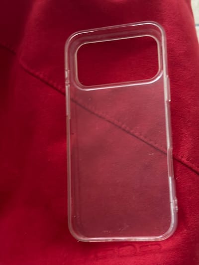 A transparent smartphone case lies on a red fabric surface. The case is rectangular with rounded corners and has a cutout at the top for a camera and sensors. The fabric surface appears to be a soft material, possibly velvet or fleece, and has a stitched seam running diagonally across the image. There are no people or animals visible in the image, nor any other objects. The lighting is diffused, casting soft shadows. The location in Crokhovo, Russia is not discernible from the image.