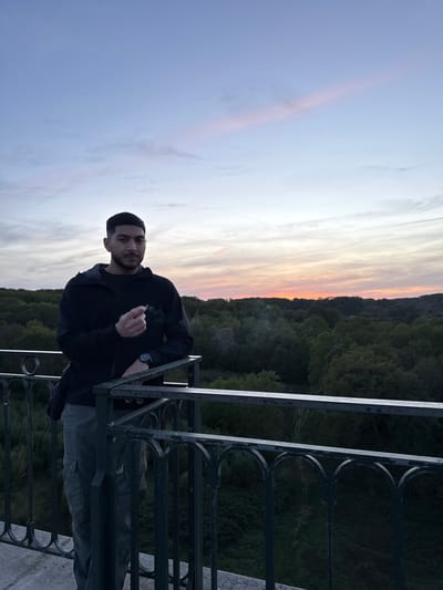 A portrait-oriented, eye-level shot captures a young man outdoors at sunset, in Gometz-le-Châtel, France. The man, with short dark hair and a beard, is positioned slightly to the left of the frame's center. He wears a dark long-sleeved top and dark pants, and a watch on his left wrist. His right hand holds a lit object, possibly a cigarette or joint, from which smoke trails upwards. He leans casually against a dark, ornate metal railing that forms the lower-right boundary of the image.
The background features a dramatic sky with soft, elongated clouds displaying warm hues of orange, pink, and pale yellow near the horizon, transitioning to light blue and grey further up. Below the horizon, a dense, dark green forest stretches across the landscape, suggesting a valley or a view from a high vantage point. The overall lighting is dim, characteristic of twilight. The man looks directly at the camera with a neutral expression. No visible text is present in the image.
