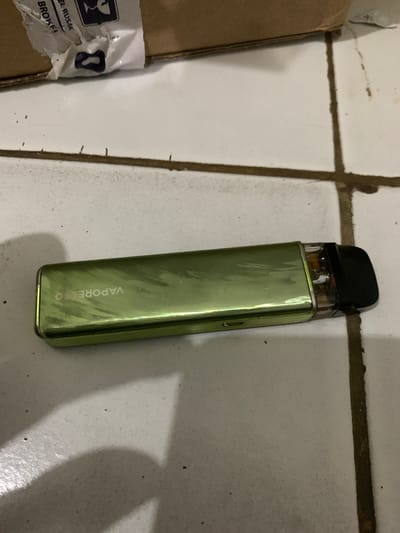 The image features a green Vaporesso vape device lying diagonally on a white tiled surface with dark, soiled grout lines. The vape has a sleek, metallic green finish, which appears somewhat scratched or worn. A black, ergonomic mouthpiece is attached to the top, above a clear pod section that shows some liquid content. The brand name "VAPORESSO" is visibly printed vertically on the side of the device.
To the left of the vape, a brown cardboard box is partially visible. A torn white shipping label on the box displays a standard "handle with care" or "fragile" symbol, along with partial text that reads "EL RUSAK" (Indonesian for "damaged" or "broken") and "BROKE". This indicates that the package may have sustained damage.
The setting is an indoor environment, likely a floor or counter, characterized by the white ceramic tiles and their dark, somewhat dirty grout. Specks of dust and dirt are visible on the tiles and within the grout lines. The lighting is even and ambient, without strong shadows, suggesting indoor artificial light or overcast daylight. No specific activity is occurring; the items are stationary, suggesting an unposed snapshot of objects in Cibinong, Indonesia.