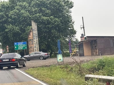 The image shows a roadside scene in the community of Nigot'i in Georgia. A black Mercedes-Benz sedan is parked on the right side of the road, its rear facing the camera. A grey sedan is parked in front of it. To the left of the road, a blue and white directional sign indicates directions to "X33353 JAPANA," "LANCHKHUTI 17," and "BATUMI 98." A large, carved stone monument stands behind the sign. On the right side of the road, there is a small wooden structure with a dark roof, possibly a shop or kiosk, with two umbrellas, one blue and one purple, displayed in front of it. A poster advertising coffee is propped up near the structure. The environment is lush and green, with tall trees and grassy verges bordering the road. The sky is overcast, suggesting a cloudy day. There are no people clearly visible in the image.