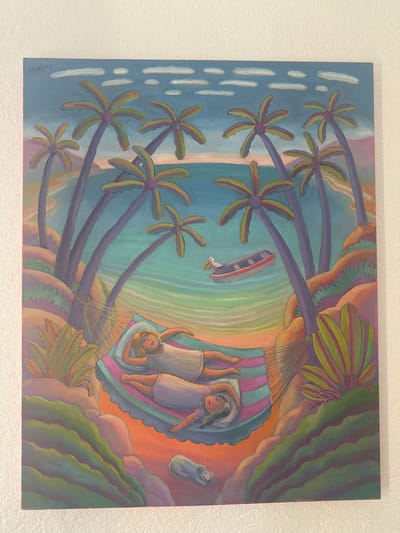 A vibrant painting depicts a tropical beach scene. Palm trees line the shore, leading to a blue sea with white ripples and a small boat accompanied by a white bird. Two girls are relaxing in a colorful hammock hanging between lush foliage. A dog lies beside the hammock. The artwork features a whimsical and slightly rotated perspective, capturing a dreamy, idyllic vacation setting. The artist's signature is visible in the lower left corner.