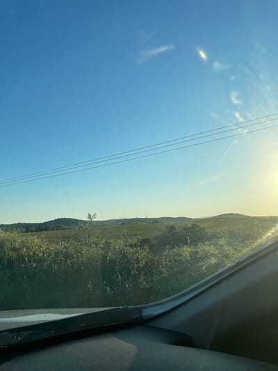 The image shows a landscape view from inside a vehicle, likely a car. The right side of the frame is dominated by the interior of the car, including part of the door and seat. Through the window, we see a rural landscape. The right side of the view depicts a grassy hill or field with vegetation. The sky is bright blue with a few wispy clouds and power lines cutting across the upper left. The sun is positioned at the top of the image, creating a bright glare. Based on the query for location in Xanthi Municipal Unit, Greece, the scene is likely a countryside road or area within Xanthi Municipal Unit in Greece, with the car moving along a road bordering a field or grassy area. The area is predominantly rural and likely has power lines running alongside the road. There are no people directly visible in the image.