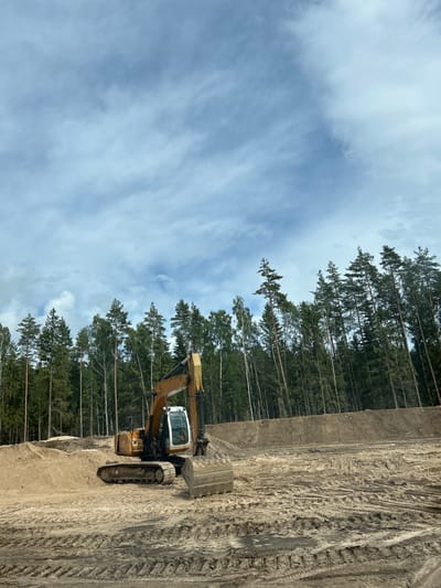 An excavator sits idle in a cleared construction site, its yellow boom and bucket prominent against a backdrop of dense green forest and a cloudy sky. The ground is disturbed earth and sand, marked with the tracks of heavy machinery. The scene appears to be in a rural or suburban area, with the forest line suggesting the edge of development.