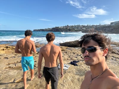 Three shirtless males are depicted on a rocky shoreline in Tamarama, Australia, under a clear blue sky with scattered cirrus clouds. The male in the immediate foreground on the right, partially cropped, has dark wavy hair, a mustache, and a goatee. He wears dark sunglasses with silver frames and a dark beaded necklace, looking directly towards the camera. To his left, two other males stand facing away from the camera, observing the ocean. The male on the far left has dark, curly hair and wears light blue swim shorts. The male in the center-right has brown, wavy hair and wears dark-colored swim shorts. Both are bare-chested. The ground consists of light brown, eroded sandstone rock formations, some with pooled water. The ocean in the mid-ground displays breaking waves near the shore, transitioning to calmer blue water further out. The distant background reveals a headland adorned with numerous residential buildings on cliffs and hills. A dark item of clothing, possibly a shirt or towel, lies on the rocks near the feet of the male in dark swim shorts. The scene is brightly lit by sunlight, casting visible shadows.