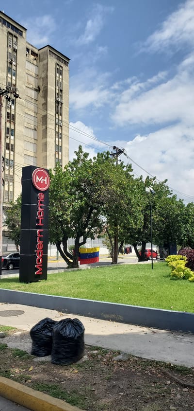This media file captures an urban street scene in Barquisimeto, Venezuela, under a bright, partly cloudy sky. The image is framed to show a commercial or residential building, a large business sign, trees, and ground features.

In the foreground, a prominent black vertical sign for "Modern Home" is visible, with the text written in red and a circular red logo featuring stylized white letters, likely "MH". To the right of the sign, on a patch of dirt and fallen leaves, are two dark black trash bags, tied and full. This dirt area borders a concrete sidewalk and a well-maintained strip of green grass.

Behind the sign and grass, several large, leafy green trees line what appears to be a street or pathway. A Venezuelan flag, characterized by its yellow, blue, and red horizontal stripes with an arc of white stars on the blue band, hangs from a pole among these trees, serving as a clear geographical identifier. Glimpses of a red vehicle and other cars can be seen in the background through the trees.

To the left, a tall, multi-story building with a light-colored, aged facade dominates the background. It features numerous windows, some with visible air conditioning units and horizontal shutters or blinds. Utility poles with a dense network of overhead wires are noticeable against the blue sky and scattered white clouds, extending across the scene and partially obscuring the building.

The scene depicts a typical daytime setting, with no people visible and no specific event or activity taking place. The overall impression is one of an active but static urban environment, characterized by commercial presence, street infrastructure, and local flora.