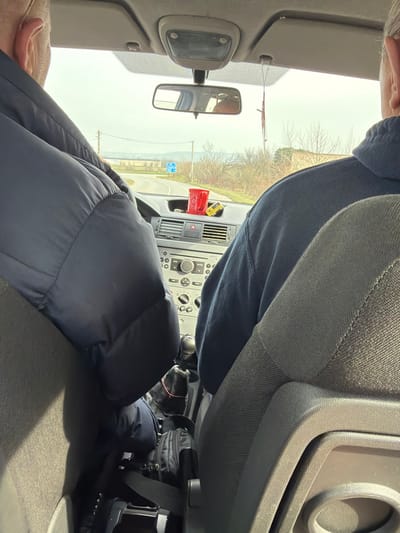The image displays the interior of a passenger vehicle, observed from a rear seat, looking forward through the windshield on a road near Kermen, Bulgaria. Two adult males are seated in the front: the driver wears a dark blue puffer jacket, and the front passenger wears a dark blue sweatshirt. On the gray dashboard, a red plastic cup and a yellow toy car are positioned. The center console includes various controls for audio and climate, with a manual gear shifter visible below. A rearview mirror is mounted, from which a small decorative pendant hangs. Through the windshield, an asphalt road curves to the left in the distance. The surrounding environment consists of bare trees, utility poles with overhead lines, and a blue informational road sign. The sky is overcast and light gray.