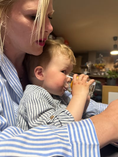 A blonde woman is lying down while holding a toddler who is drinking from a glass. Both are wearing striped shirts. The exact location in Baillonville, Belgium is not discernible from the image.