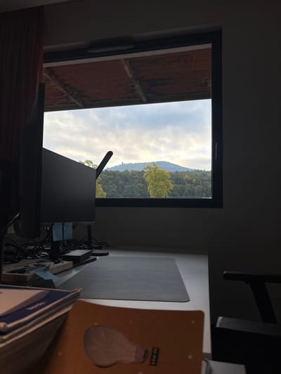 The image displays an indoor workspace with a direct view through a window to an outdoor natural landscape in Lièpvre, France. In the foreground, a white desk occupies the lower half of the frame. A dark computer monitor, displaying a dark screen, is positioned on the desk to the left, with a black desk lamp featuring a curved arm situated behind it. Clustered cables are visible behind the monitor. Several small blue sticky notes are attached to the monitor stand and other items on the desk. A rectangular grey desk mat is centered on the desk. A stack of papers or books is partially visible in the lower-left corner, and a light-brown notebook with a stylized lightbulb graphic is in the lower-right foreground. A section of a dark office chair's armrest and back are visible on the far right.

Through the dark-framed window, an overcast sky with grey clouds extends above a forested mountain or hill range. Dense green foliage covers the landscape, with one tree in the mid-ground exhibiting lighter green or yellowish leaves. Above the window frame, a partial view of an exterior overhang with reddish-brown wooden planks is discernible. A reddish-brown curtain is visible on the left side of the window. No individuals or animals are present in the scene.