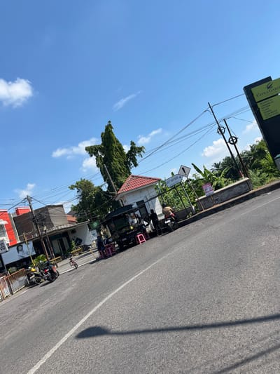 A low-angle, full outdoor shot captures a street scene on a sunny day in Entalsewu, Indonesia. A paved road curves through the foreground, with a white line marking the lane. To the left, a white and red building is visible, with a sign reading "GIGI". Further down the street, a food cart is parked, with people gathered around it. A motorbike is parked in front of the food cart, and another motorbike is parked to the left. In the background, a tall green tree stands out against a bright blue sky with scattered white clouds. A large black sign with yellow lettering, reading "CITRA GARDEN" and listing various amenities, is positioned on the right side of the image. Utility poles with numerous wires are visible throughout the scene, indicating an urban or suburban setting. The overall impression is one of a bustling street in a tropical climate.