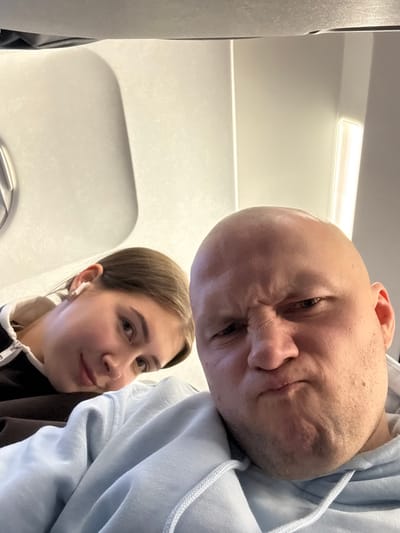 The image depicts two individuals inside an aircraft cabin, likely at Емельяново Airport, Russia, given the provided context.
The foreground is dominated by a bald adult male on the right, wearing a light blue hooded sweatshirt. He is captured in a close-up, making an exaggerated, comedic grimace; his eyebrows are furrowed, eyes squinted, nose wrinkled, and lips pursed downwards. His gaze is directed towards the camera.
Behind and to the left of the male is a young adult female. She has light brown hair and is wearing a dark-colored top with a white inner collar, possibly a zipped jacket or hoodie. A white wireless earbud is visible in her left ear. Her head is tilted, resting on a dark surface, and she displays a slight, knowing smile, looking towards the camera or the male.
The background shows the light-colored, curved interior panels of an aircraft cabin, consistent with overhead compartments and ceiling structures. A vertical light source or window frame is partially visible in the upper right quadrant. The scene suggests a selfie taken by one of the individuals.