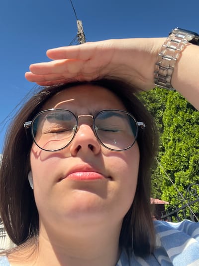 This is a close-up, rotated selfie of a young woman outdoors. She has dark hair and is wearing round, wire-rimmed glasses and a silver-toned watch on her left wrist. Her eyes are closed, and her lips are slightly parted and appear to have lip gloss on them, suggesting a relaxed or contented emotion. Her right arm is raised and bent, with her hand resting on her forehead, shielding her eyes from the sun. The background shows a clear blue sky, lush green foliage, and a glimpse of a building with a striped awning. The lighting suggests it is daytime, possibly late morning or afternoon, with bright sunlight. There is no visible text in the image.