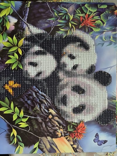 This media file is a diamond painting depicting a family of three pandas nestled in a lush, green environment with vibrant red flowers and a blue butterfly. The pandas, represented by a mosaic of small, shimmering gems, appear to be a mother and her two cubs. The mother panda is larger, with her cubs snuggled close. One cub is peeking out from behind its mother, while the other is positioned in front. The background features a rich, dark green foliage with hints of purple, suggesting a dense jungle or forest setting. The red flowers add a pop of color against the greenery and the pandas' black and white fur. A delicate blue butterfly flutters in the upper right corner of the painting, adding a touch of whimsical movement to the scene. The lighting appears to be soft and diffused, possibly indicating a gentle daytime or twilight ambiance. The overall impression is one of peaceful family bonding within a serene natural habitat. There is no visible text in the image.