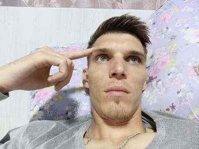 A medium shot captures a young adult male lying on a floral-patterned pillow. The man has light skin, short brown hair styled upwards, and blue eyes. His right index finger is touching his forehead, and he is looking upwards. He has a short goatee and is wearing a striped shirt and a silver chain necklace. The background includes a textured wall and the floral pillow. The setting is likely a bedroom in Uchquduq, Uzbekistan.
