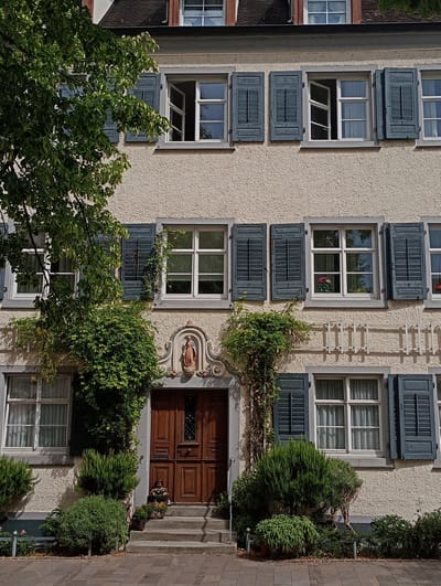 The image shows the facade of a building in Meersburg, Germany, with a light-colored, stucco exterior and dark blue shutters on multiple windows. The building has several floors, with a prominent wooden door at ground level, flanked by steps and greenery. A small statue of a person is positioned in an alcove above the doorway. Vines and potted plants are visible around the entrance and on the facade. Sunlight casts shadows, suggesting it is daytime.