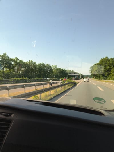 The image captures a view from inside a vehicle traveling on a highway in Marl, Germany. The foreground is dominated by the dark dashboard and windshield of the car. The windshield has some dust or water spots, obscuring the view slightly.

The highway is multi-lane, with other vehicles visible in the distance. A white car is in front, with traffic continuing ahead. On the left side of the highway, there is a guardrail and a grassy embankment lined with trees. The trees form a dense green line along the side of the road. On the right side, more trees are visible.

The sky is a clear, bright blue, indicating it is daytime, likely morning or afternoon. The sun is casting shadows, suggesting it's not midday. The overall impression is of a sunny day. A green sticker with white text and a symbol is visible on the right side of the windshield. The setting is a typical German autobahn, given the highway design and the location context provided. There are no people clearly visible in the image, only the interior of the vehicle and the passing scenery. The activity taking place is travel on the highway.