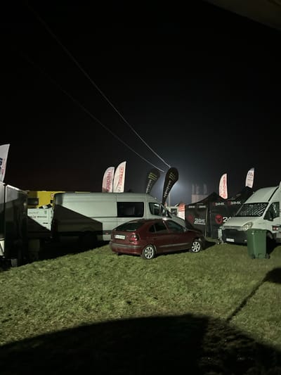 This image depicts a nighttime scene in a grassy field in Poroy, Bulgaria, illuminated by artificial light. A dark red hatchback car, with Bulgarian license plate A 0353 BA, is parked diagonally in the center-left foreground. To its left is a light-colored cargo van, partially visible. To the right of the red car, a white cargo van is parked, facing right, with a green waste bin positioned in front of it. In the background, multiple tall, slender advertising flags are visible, some featuring "MONSTER" branding, others with "MOTO" and Cyrillic text ("АВТОСЕР," "ДАРАН," and others). Dark canopy tents with "24MX" branding are situated behind the vehicles. Overhead, thin lines, likely power or guide cables, traverse the dark sky. A strong artificial light source in the background creates a hazy glow. No individuals are visible in the scene.