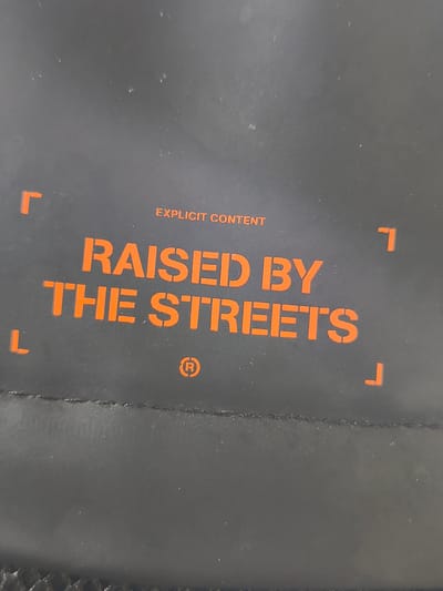 The image is a close-up, vertically oriented photograph of a black surface, likely a bag or fabric, with orange stencil-like lettering. The text is centered and reads "RAISED BY THE STREETS" on two lines. Above this, centered, is the phrase "EXPLICIT CONTENT". To the left of "RAISED BY", there are two orange square brackets facing inwards, forming a "Γ" shape. To the right of "STREETS", there are also two orange square brackets forming a "Γ" shape. Below "STREETS", a small orange registered trademark symbol "®" is visible. The surface appears to have a subtle texture and some minor dust or imperfections. The lighting suggests an indoor setting. The image does not contain any elements that place it in Momchilovtsi, Bulgaria, or any other specific location.