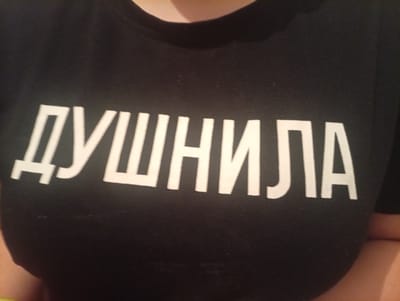 The media file shows a close-up, chest-level shot of a person wearing a black t-shirt with white text printed on it. The text is in Cyrillic script and reads "ДУШНИЛА". The person's neck and upper chest are visible, suggesting a casual setting. The lighting appears to be indoor, possibly artificial, given the absence of shadows suggesting natural light. The focus is on the t-shirt and the text. There are no other discernible objects or elements in the immediate background or foreground. The overall impression is a simple, close-up photograph of a piece of clothing with a message. The location context is Shagonar, Russia.