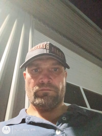 A forward-facing photograph depicts an adult male from the chest up, positioned against a structure featuring light-colored, vertical corrugated metal siding under low light conditions, consistent with nighttime or an indoor setting. The male subject has fair skin, a short brown beard and mustache, and blue eyes, maintaining a neutral expression while looking directly at the camera. He wears a dark baseball cap with a light-colored, partially obscured logo on the front brim, and a dark blue collared polo shirt with three white buttons, the top two of which are unbuttoned. A window-like or framed structural element is visible in the upper right background. The image carries a "shot by motor g - 2025" watermark in the lower left corner. The specific location within the city of Salem, United States, cannot be determined from the visual information provided.