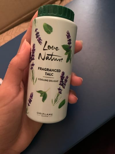 A human hand grips a white cylindrical bottle of talcum powder, identified as "Love Nature FRAGRANCED TALC COOLING DELIGHT" by "ORIFLAME SWEDEN." The bottle has a green, ribbed screw-top cap and is adorned with illustrations of purple lavender sprigs and green leaves. The hand displays a light to medium skin tone; visible fingernails are clean and rounded. The background is out of focus, presenting a dark blue planar surface on the right and a lighter, textured beige or brown surface on the left. The specific location in the city of Skhirat, Morocco, cannot be determined from the visual information provided.