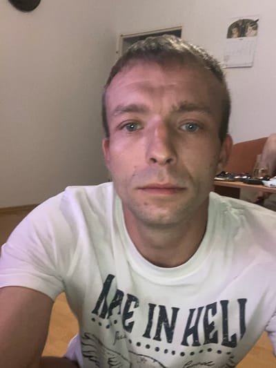 The image is a close-up selfie of a man with fair skin, short brown hair, and blue eyes. He is wearing a white t-shirt with black lettering that appears to read "ARE IN HELL". The background is a plain, light-colored wall and a wooden floor. There is also a hint of an orange chair and a desk with objects on it in the background, suggesting an indoor setting, possibly a home or an office. No specific location in Košice, Slovakia, can be identified from the image.