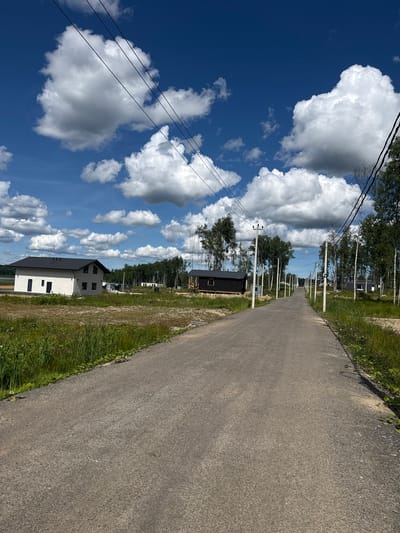 The image depicts a rural road leading through a developing residential area under a bright blue sky dotted with fluffy white clouds. The road is paved and appears to be a two-lane street. On either side of the road, there are power lines supported by white poles. Several modern houses, some appearing to be under construction, are scattered throughout the landscape. These houses are a mix of architectural styles, with some featuring white facades and dark roofs, while others are smaller, dark-colored structures. Tall, slender trees, predominantly birches, are interspersed among the houses and line the sides of the road. The ground is covered in dry grass and dirt, indicating a natural, undeveloped environment. In the distance, a body of water can be seen to the left, and a dense forest borders the area to the right. The scene suggests a quiet, suburban or rural setting, possibly a new development on the outskirts of a town. The specific location in Zashchita, Russia is not visually evident from the image itself.