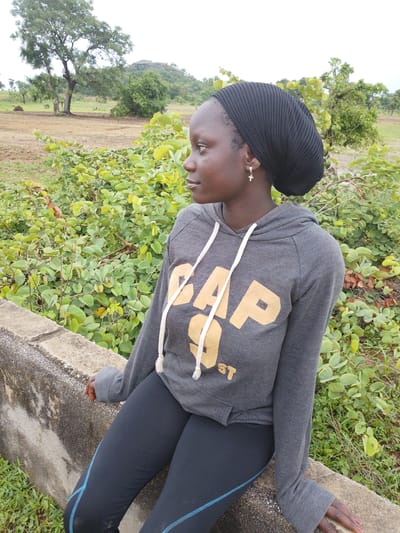 In a medium shot, a young woman wearing a black knit cap and a grey Gap hoodie with gold lettering sits on a concrete ledge. She is facing to her left, with her head turned slightly to reveal her profile. She has a small gold cross earring in her left ear. The background consists of a lush green bush, a brown dirt field, and trees under an overcast sky. The location is the city of Jauro Garmaho, Nigeria.